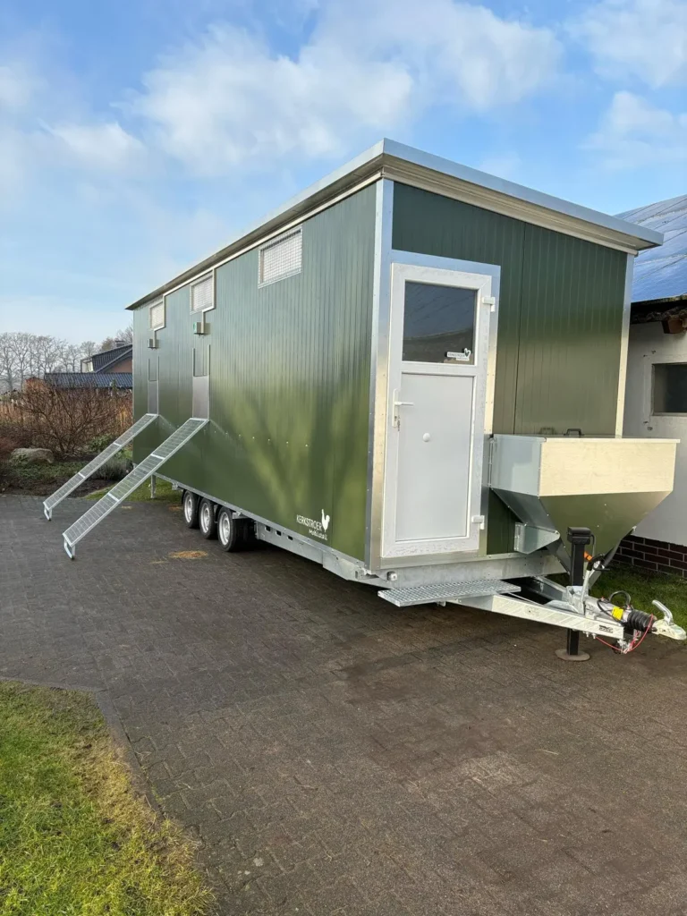 Ein mobiler Stall in grünem Design mit Rampen, einem weißen Eingang und einem Futtersilo, steht auf einem gepflasterten Gelände unter einem blauen Himmel.